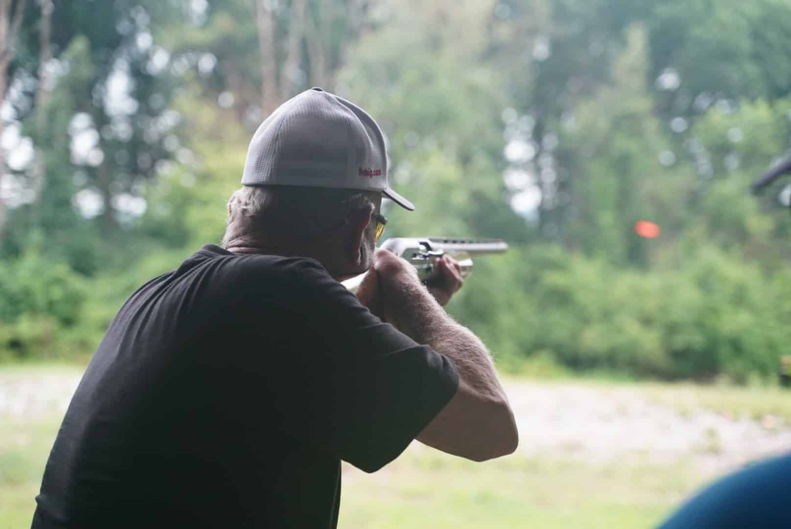 A close-up, side-view shot of a person wearing a grey cap and a black shirt. They are holding a shotgun to their shoulder, aiming. An orange clay target is flying in the distance. The person is in a wooded area with trees in the background.