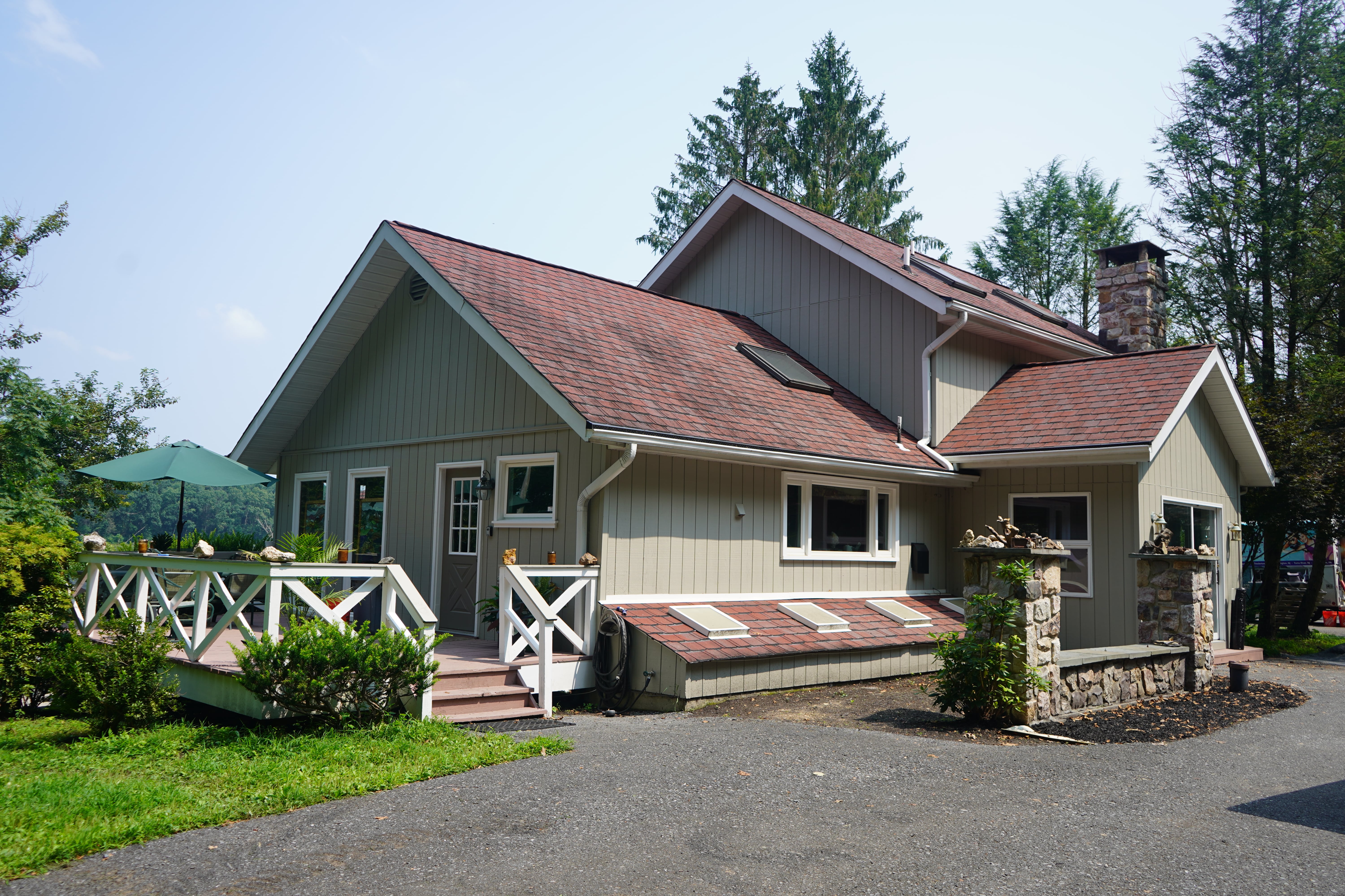 A tan house with a red roof, featuring a chimney made of stone and a large wooden deck with a white railing. The deck overlooks a grassy field with a line of trees in the background. The house is surrounded by a driveway and bare trees, and the ground is covered with fallen leaves.